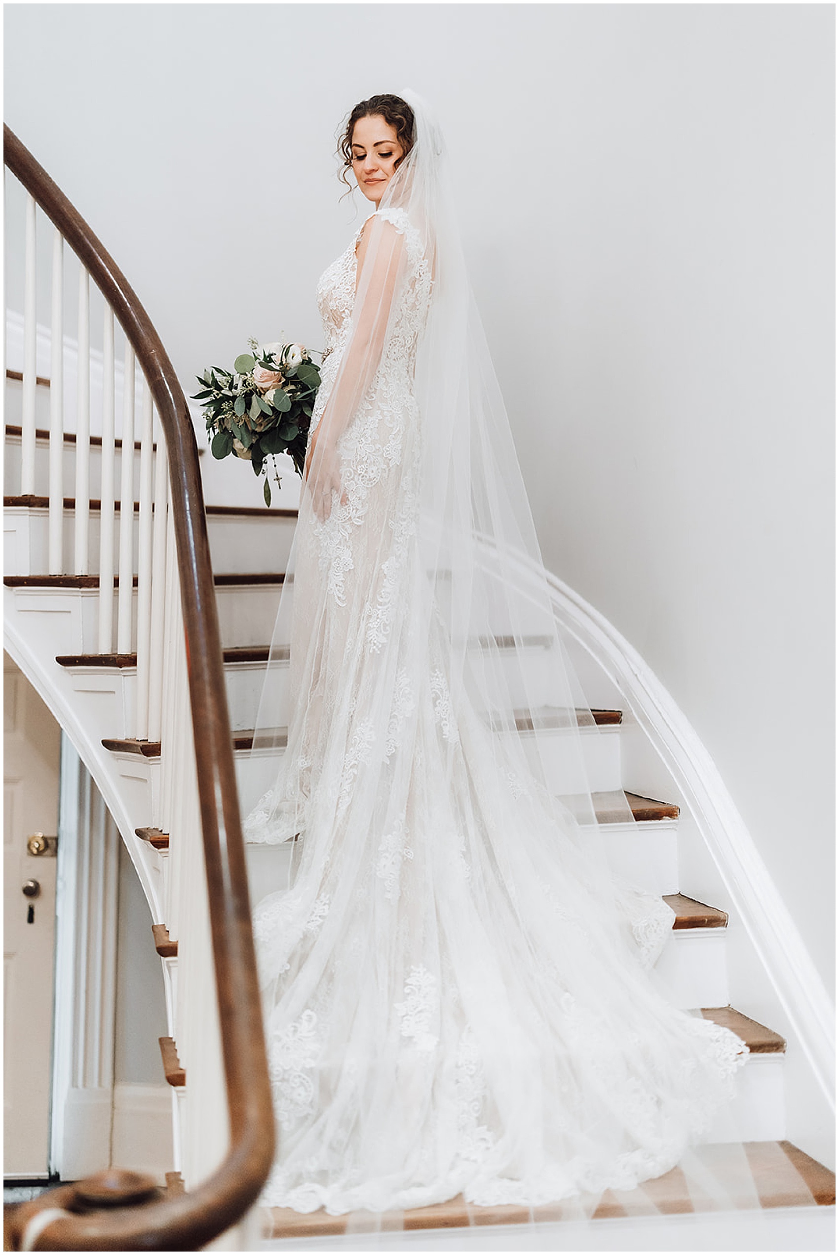 Bride will extra long train and veil stands in a stairwell with dress flowing down stairs