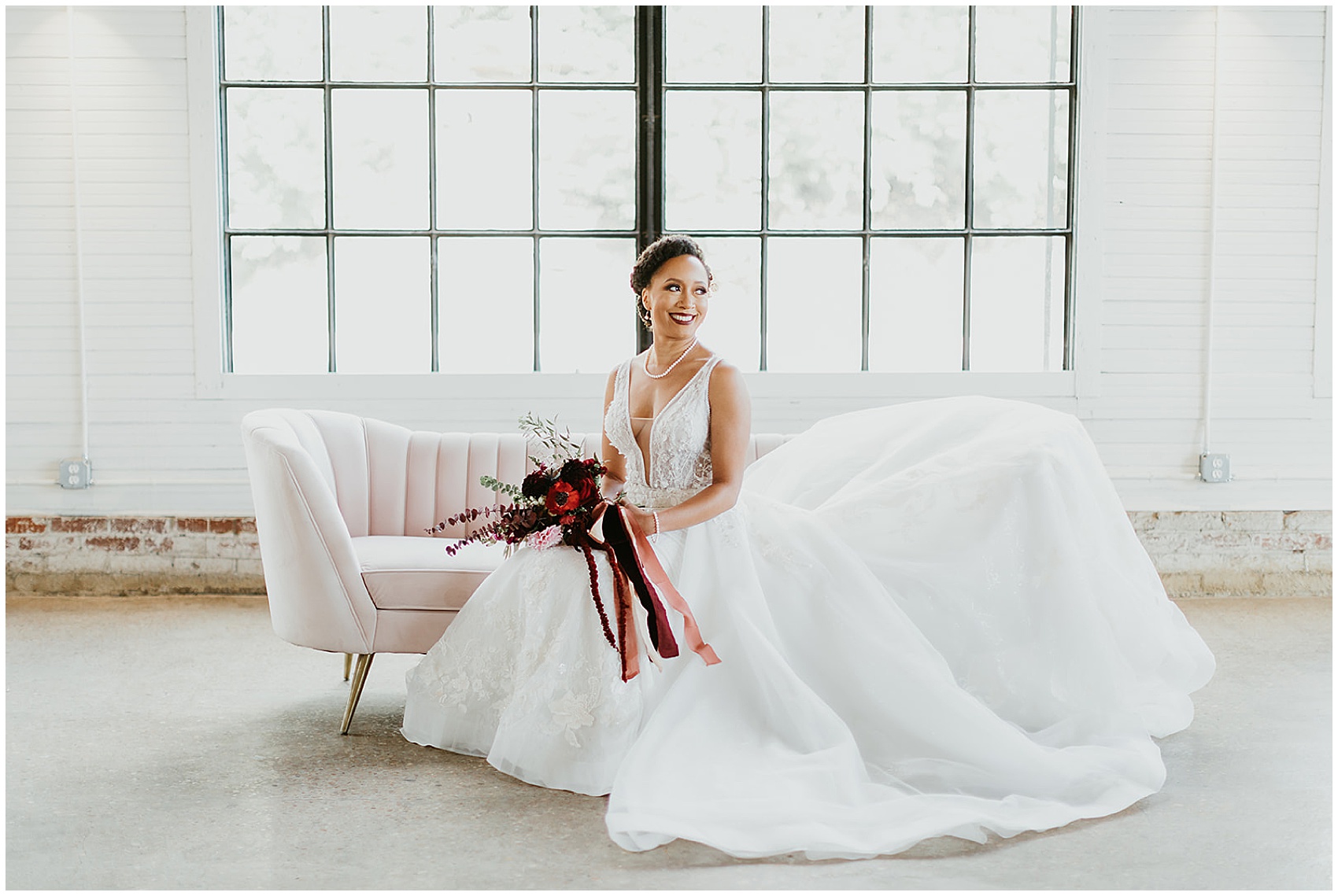 Bride in large white dress sits on a couch in front of a large window holding her red bouquet PLS Coordinate