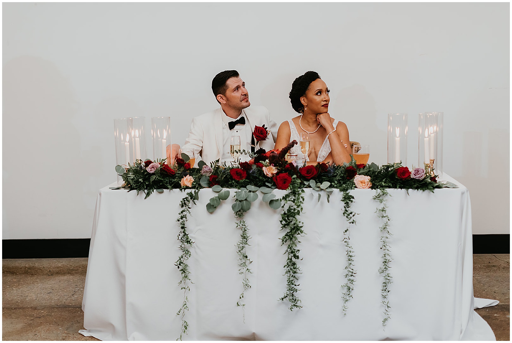 Newlyweds sit on their decorated head table with candles and roses listening to speeches PLS Coordinate