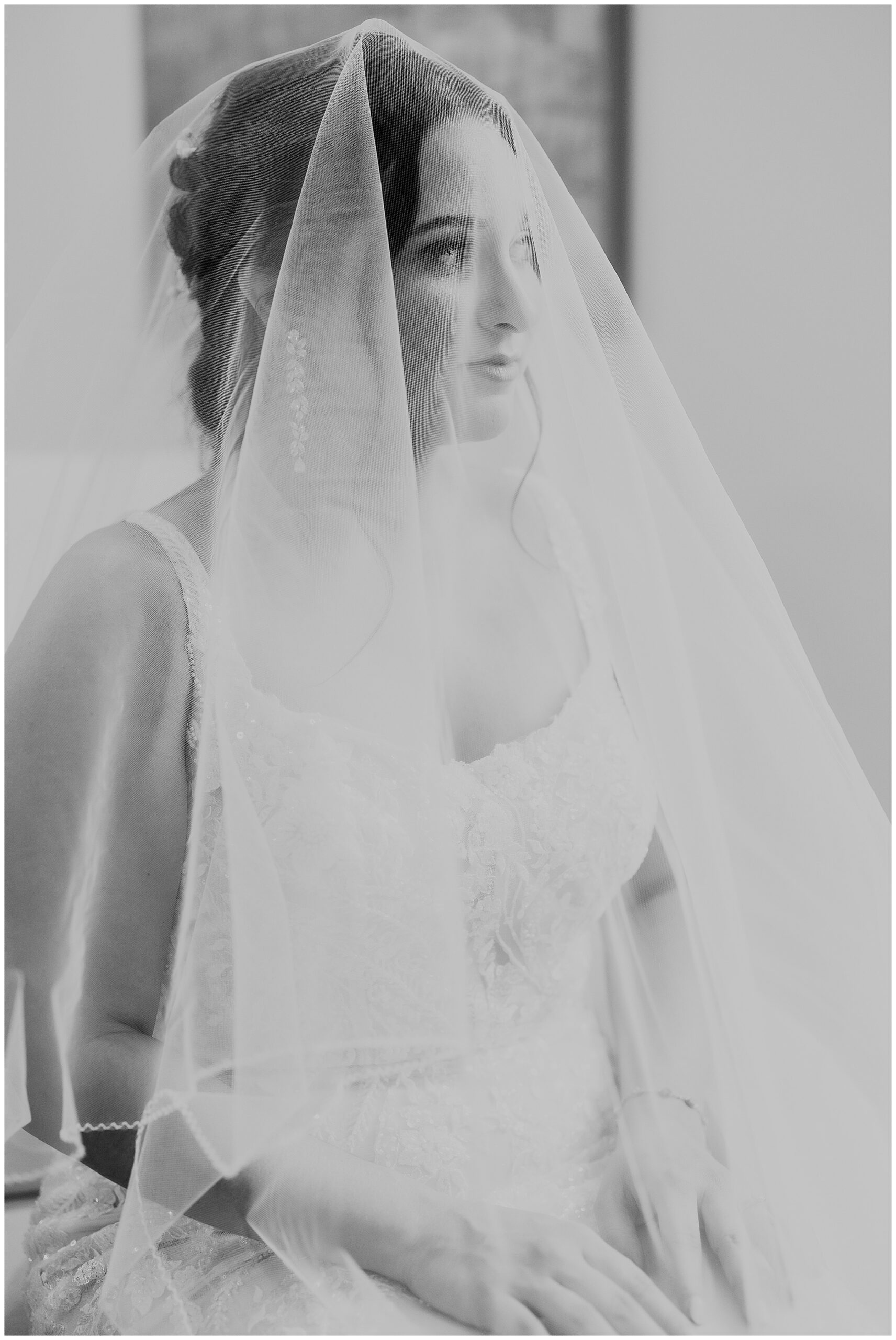 Bride in a white lace dress sits in a room with her veil over her face