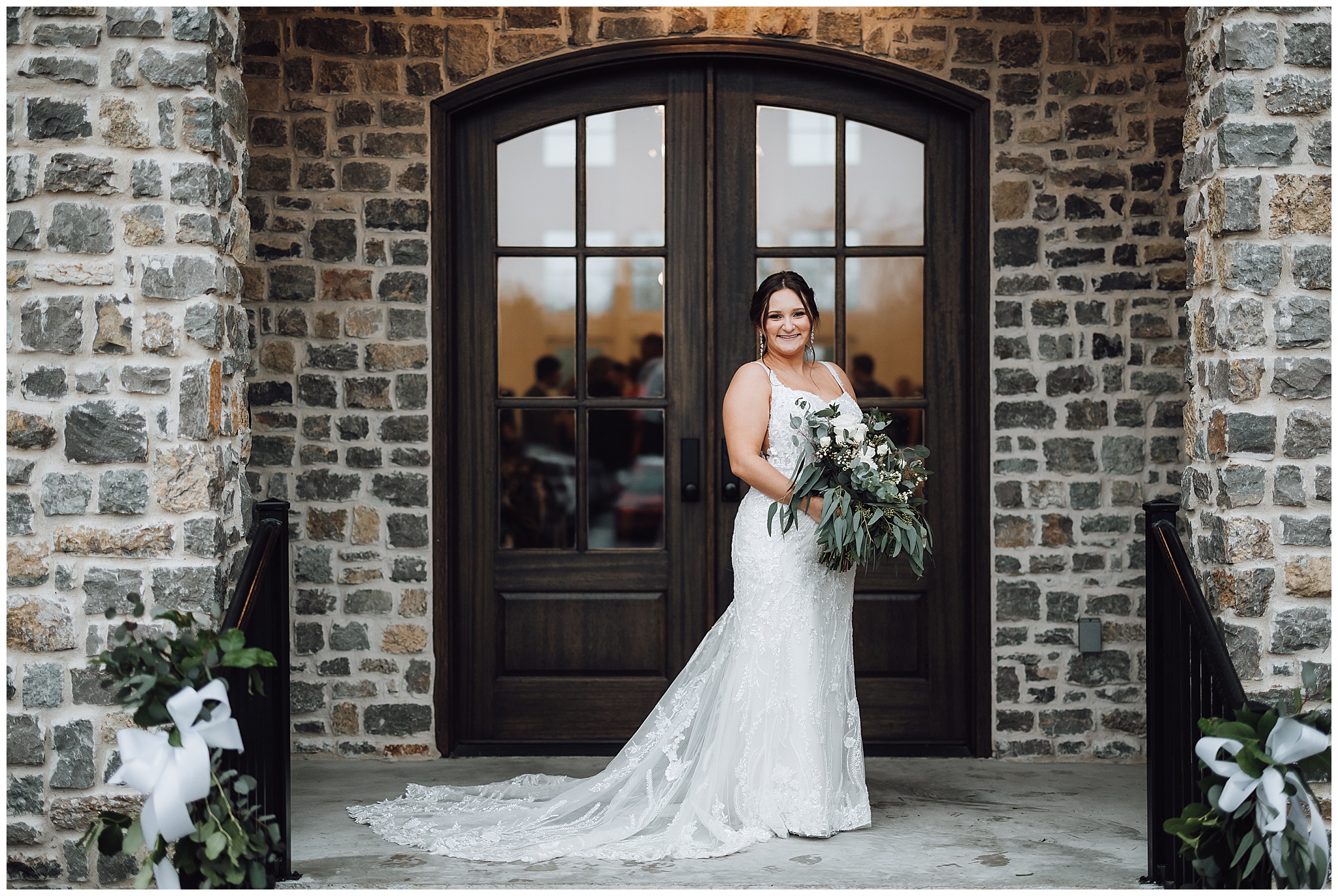 Bride stands in front of the entrance to her reception in front of large dark wooden doors and stone wall and columns of The Venue at Birchwood