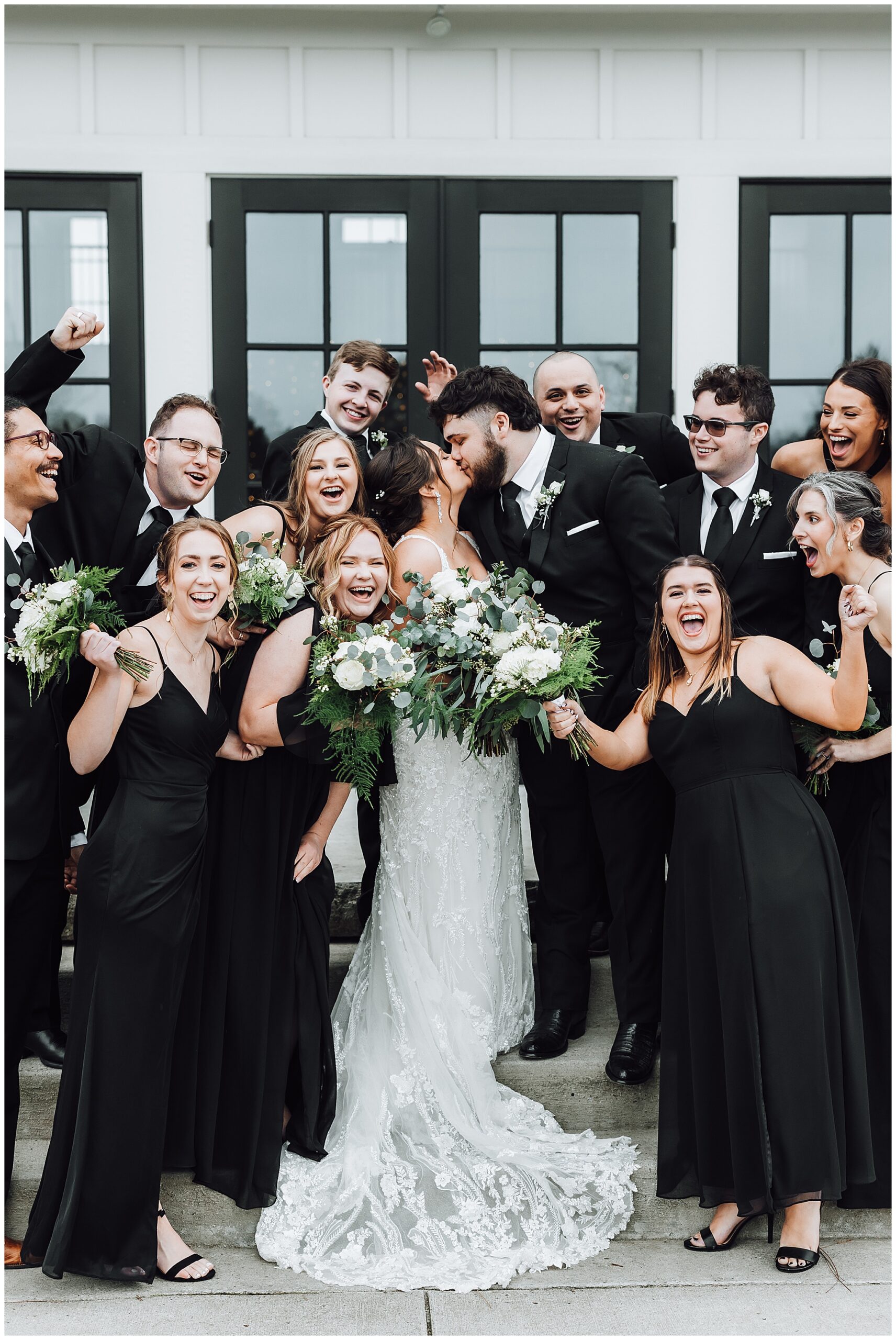The whole wedding party celebrates as the bride and groom kiss on stairs