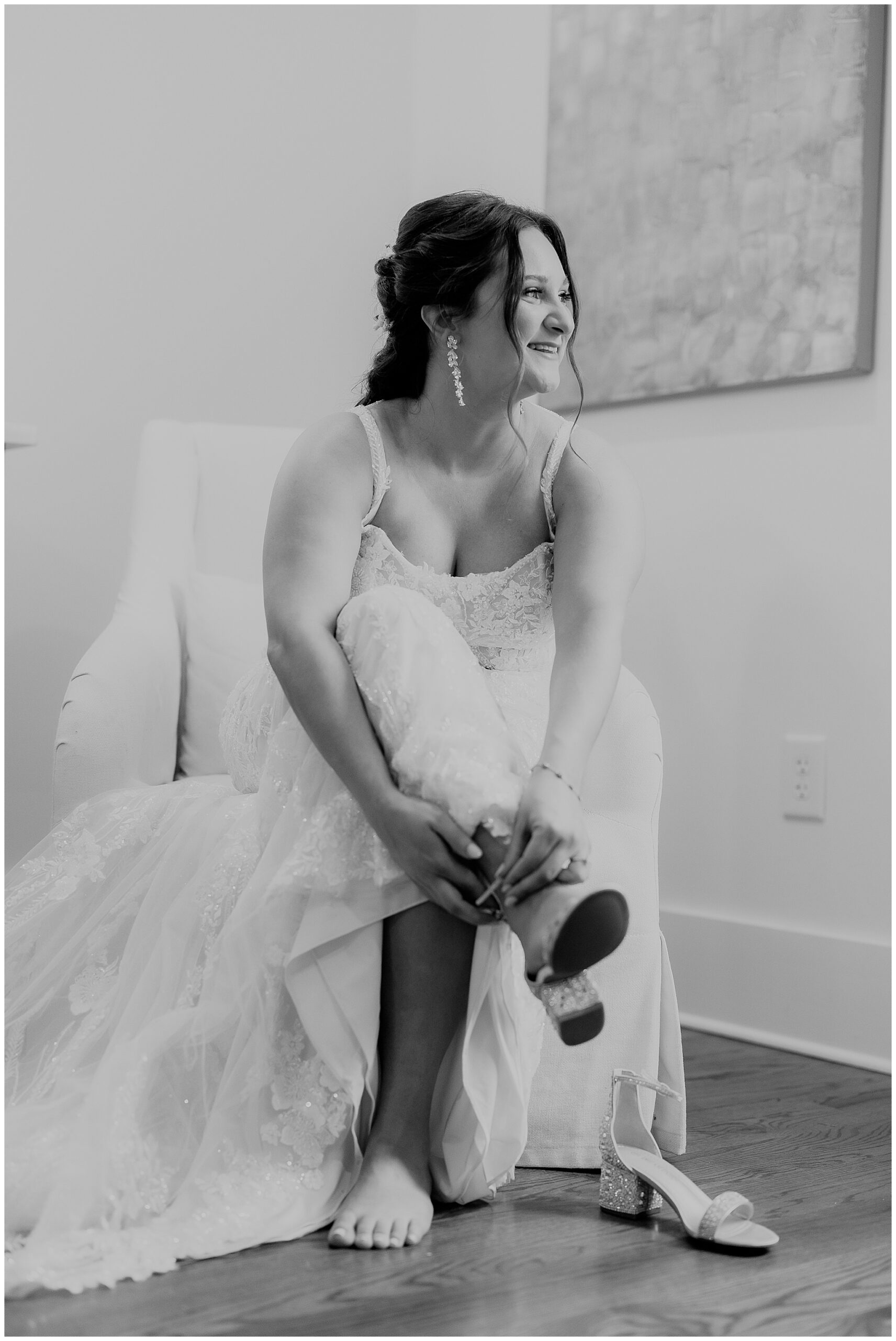 Bride in a white lace dress sits on a white chair while strapping on her diamond-encrusted heels