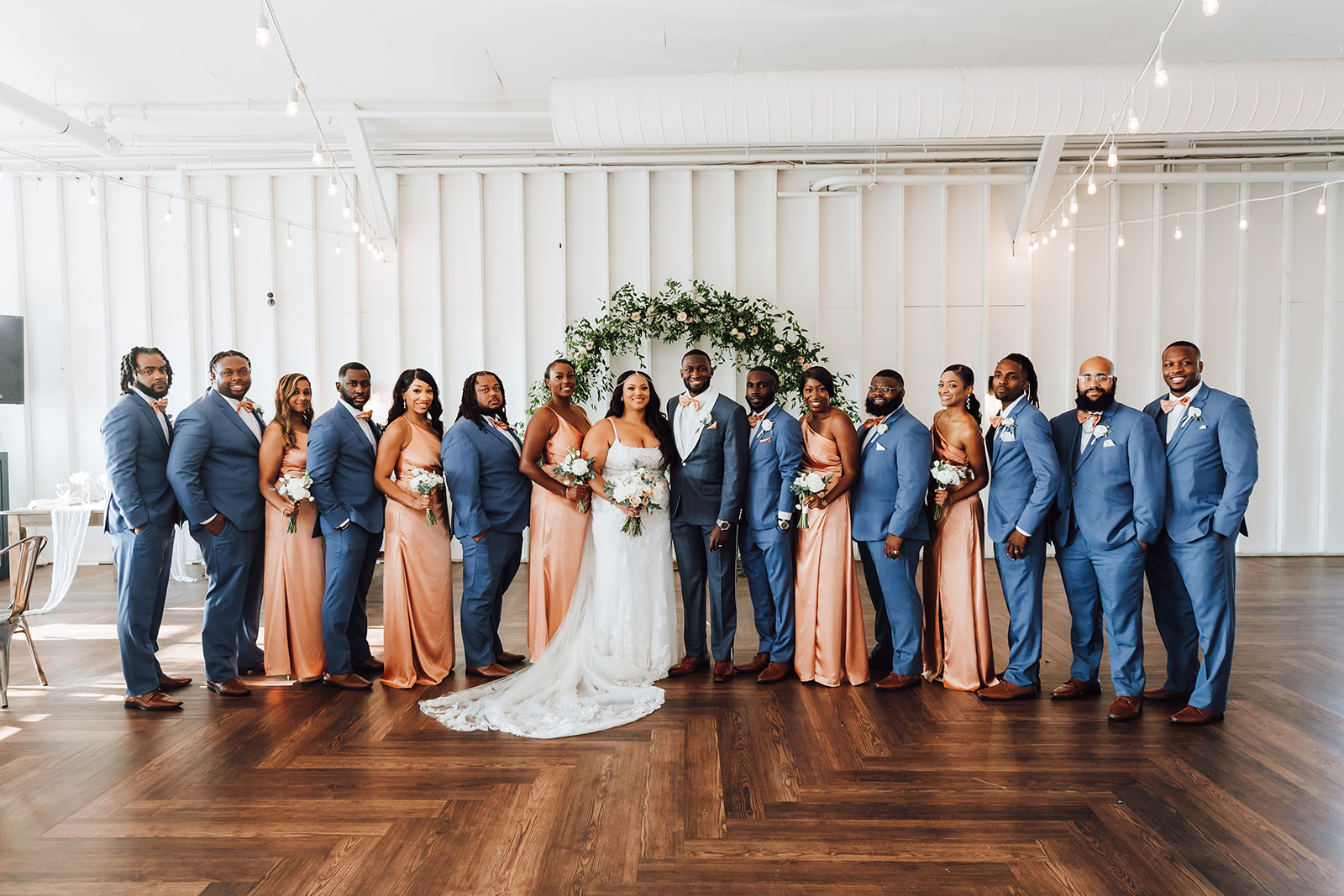 A large wedding party stands together in blue suits and pink dresses