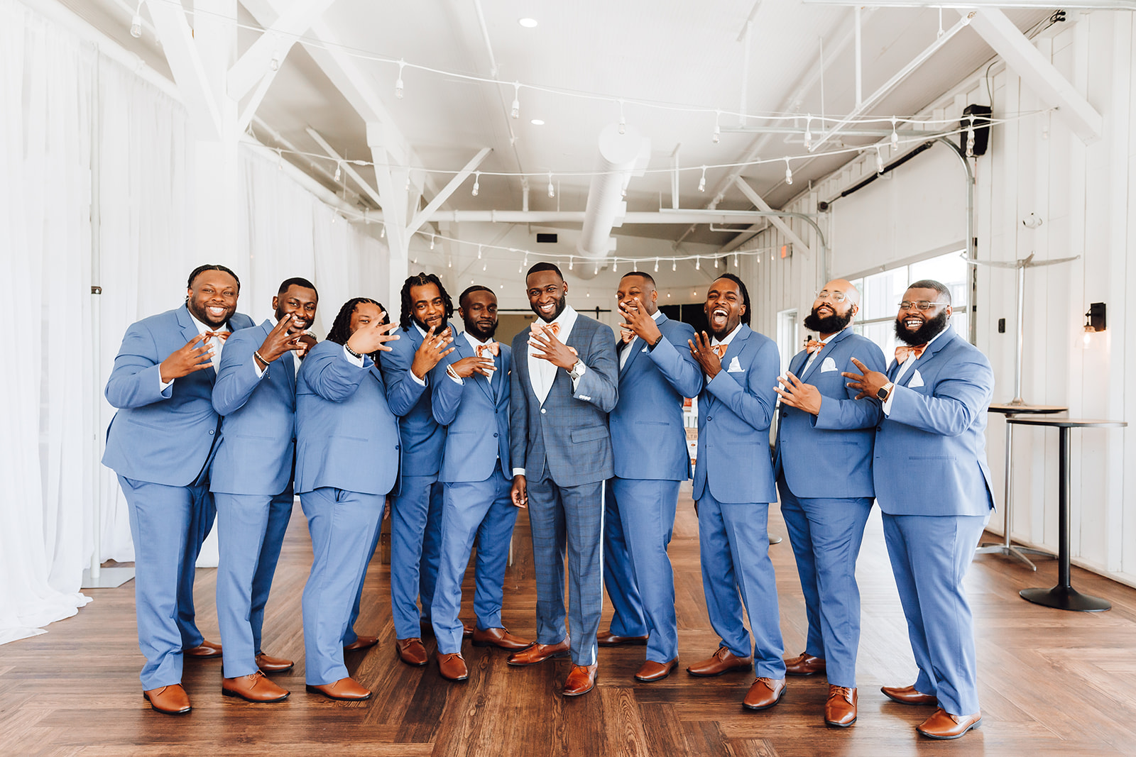 A groom and his wedding party put up four fingers while standing in a reception room 14Tenn Wedding