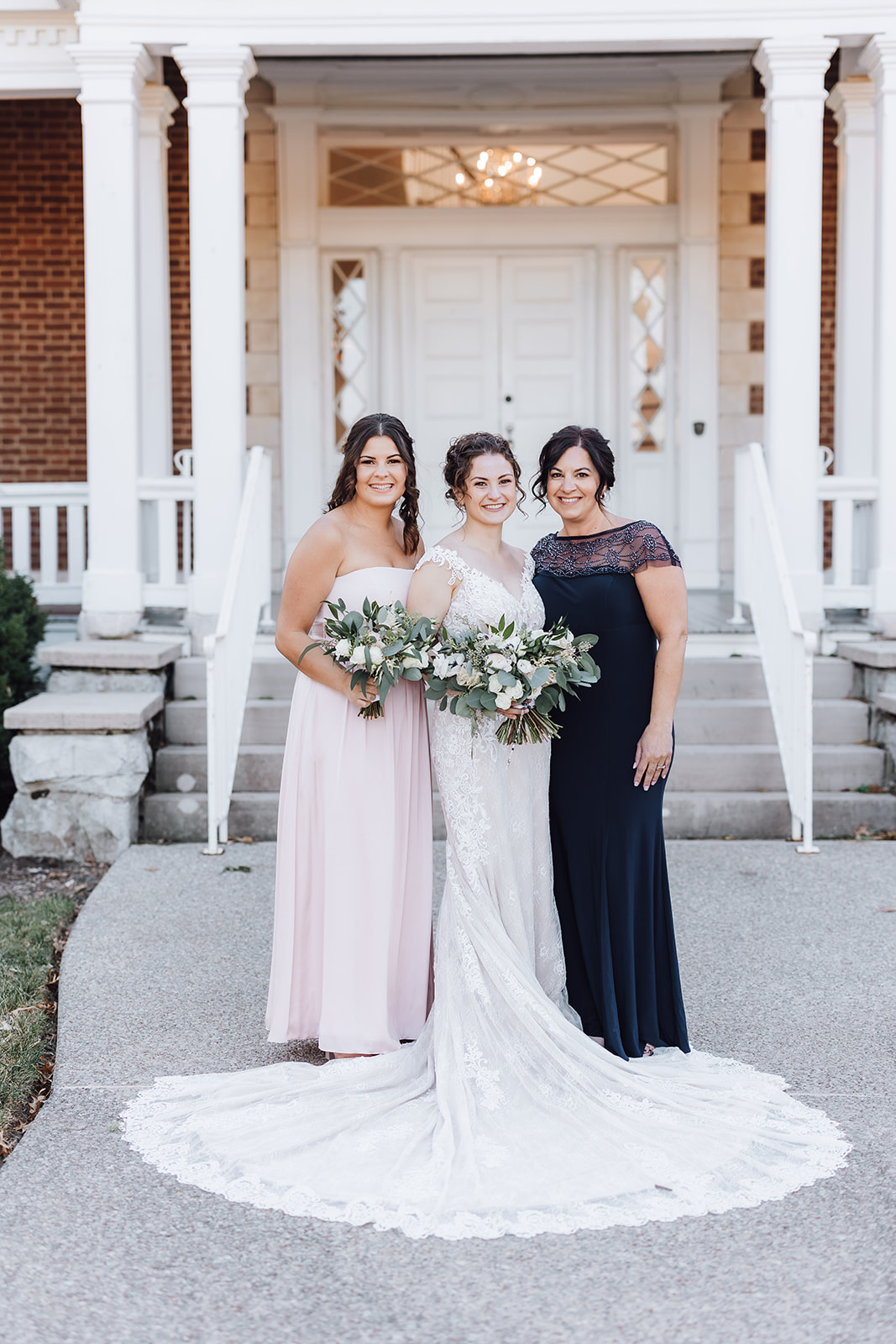 A bride in a white lace dress with a long round train stands with her mother and sister in front of the Ravenswood Mansion