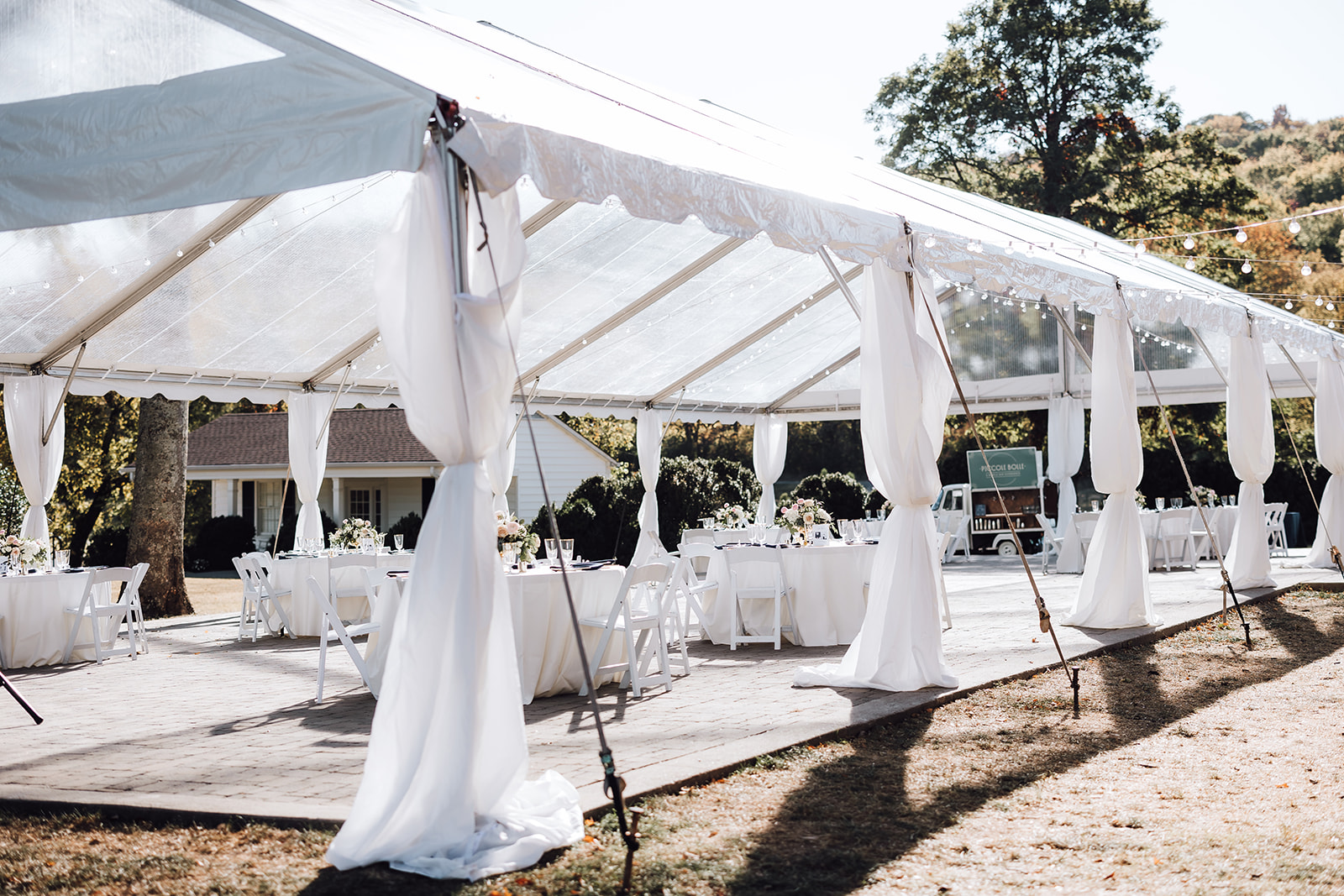 A wedding reception set up outside in a white tent with market lights and white linens Ravenswood Mansion