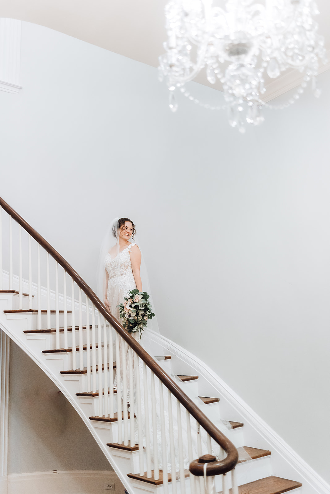 A bride looks over her shoulder while walking up a large curved wooden staircase in a lace dress Ravenswood Mansion
