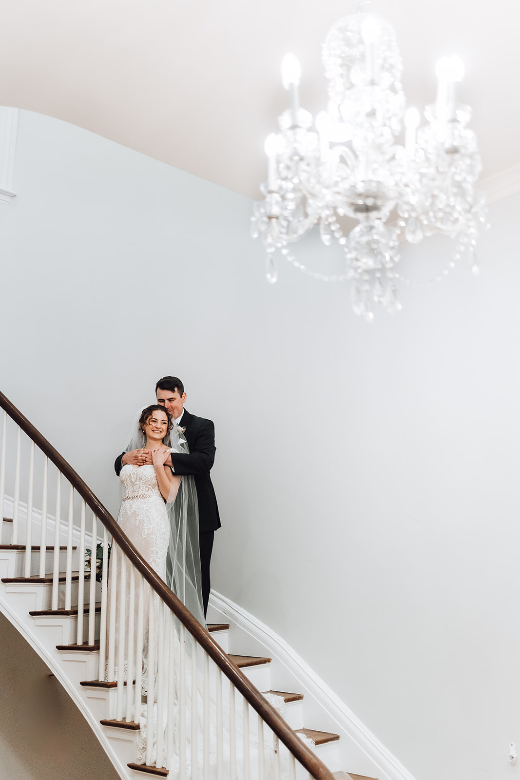 A bride and groom hug and snuggle on a large wooden staircase under a crystal chandelier Ravenswood Mansion